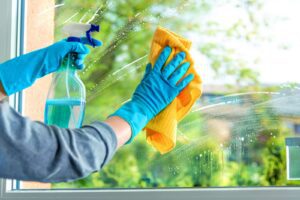 Two hands wearing blue latex gloves spraying cleaning solution onto a window.